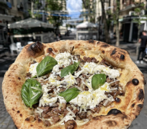 La pizza di Giuseppe Vesi non è soltanto una specialità da assaporare, ma una storia da leggere tra i profumi del forno, la memoria dei vicoli e il respiro della terra. Oggi, in via Luca Giordano 162, al Vomero, la sua «Trattoria Pizzeria» trova dimora tra gli alberi superstiti dell'antica collina, quasi a voler legare il destino di un uomo alle radici di un territorio e al raggiungimento delle vette più alte. Per comprendere il cammino di Giuseppe bisogna però risalire tre generazioni. Nato nella Sanità, riceve dalla famiglia paterna l'eredità delle botteghe, dove il nonno lavorava come pizzaiolo, in un'epoca nella quale il mestiere era considerato umile e il posto fisso rappresentava l'ambizione più sicura. Il padre, pur scegliendo un impiego stabile, continuava a preparare pizze nei fine settimana, portando con sé il figlio tra i forni del centro storico. Dalla famiglia materna, invece, Giuseppe eredita il rapporto con la campagna: un terreno collinare alle spalle dell'attuale metropolitana del Frullone, tra ciliegi e animali da cortile, dove apprende il valore della materia prima genuina. A vent'anni apre la prima pizzeria in via Bellini, seguita da quelle di via Tribunali e San Biagio dei Librai, avviate con i fratelli, ancora oggi alla guida degli indirizzi storici. Ma il tempo, per chi osserva il proprio mestiere con inquietudine creativa, non rimane mai immobile. La nascita della pizza gourmet: un'idea coniata e registrata da Vesi Circa quindici anni fa, le pizze classiche iniziano a non bastargli più. Margherita, marinara, ripieni e quattro formaggi cedono il passo alla ricerca di nuovi equilibri. Nasce così, a viale Michelangelo, «Giuseppe Vesi Pizza Gourmet & Aperitif», un progetto nel quale prende forma anche l'espressione «pizza gourmet», coniata da Giuseppe oltre vent'anni fa e successivamente registrata come marchio. Una scelta destinata a suscitare discussioni, poiché innovare la tradizione napoletana significa spesso affrontare lo sguardo severo di chi considera ogni cambiamento una rinuncia al passato. Una trasmissione televisiva dedicata proprio al confronto tra tradizione e innovazione porta Vesi all'attenzione nazionale, suscitando interesse anche tra operatori giunti dall'estero per comprendere quella nuova direzione. Dietro il nome, tuttavia, non vi è una semplice operazione commerciale, ma oltre vent'anni di studio: farine da grani italiani biologici, varietà antiche, biga, poolish, lievito madre e prefermenti, in alternativa all'impasto diretto allora prevalente. Un percorso che oggi molti rivendicano di aver seguito per primi, ma che solo Giuseppe può datare con certezza, avendo per primo coniato l'espressione e registrato il marchio. Una ricerca sviluppata anche attraverso la collaborazione con Slow Food, scegliendo oli da cultivar di nicchia, conciato romano, pomodorino del piennolo, all'epoca Presidio, e San Marzano acquistato direttamente dai produttori. Una filiera corta nella quale il grano italiano biologico rappresenta una scelta di qualità, anche quando comporta un costo maggiore per il consumatore. Da Napoli a Roma e Milano: l'espansione del marchio Vesi Il progetto approda poi in via Luca Giordano, dove la «Giuseppe Vesi Trattoria Pizzeria» unisce pizza, cucina e aperitivo. La proposta si estende anche a Roma, con «Vulcano» a Ponte Milvio, dedicato alla cucina romana, e a Milano, con la «Locanda degli Arcimboldi», dove convivono pizza e gastronomia napoletana. La pizza al Vomero: menu, gusti e proposta gastronomica Al Vomero, la pizza si presenta in un disco di circa 280 grammi, sottile, dal bordo appena accennato e dalla grande scioglievolezza, tanto ampio da debordare dal piatto. Accanto alla Margherita con provola, San Marzano DOP, provola a lenta maturazione, Parmigiano Reggiano DOP e basilico, arrivano le Special Gourmet e i ripieni, tra cui quello alla scarola con fiordilatte di Agerola, capperi, olive nere e pinoli. Una carta di circa trenta gusti, completata da montanare, crocché, fiori di zucca e frittatine. La vicinanza al museo di San Martino e la presenza di numerosi B&B rendono il Vomero un crocevia tra residenti e turisti: capitava così che i gruppi in visita, cercando anche un primo piatto oltre alla pizza, sfuggissero al locale per mancanza di alternative. Da qui la decisione di ampliare l'offerta con la cucina dello chef aversano Luigi Gerbino: pasta e patate con la provola, gnocchi alla sorrentina, parmigiana di melanzane, genovese, diventata un piccolo caso sui social, scarpariello, polpette al pomodoro, filetti, tagliate, galletto e parmigiano. Un investimento che, a un anno di distanza, si è rivelato decisamente vincente. L'area bar propone pizza fritta e una carta di circa sessanta gin, abbinati a cinque o sei toniche. Un'organizzazione affidata a professionisti, tra direttore-barman e chef dedicato, consente a Giuseppe di concentrarsi sulla pizza e sul rinnovamento stagionale del menu. Pizza senza glutine a Napoli: il primato della famiglia Vesi La storia della famiglia comprende anche un importante capitolo dedicato al senza glutine. Nel locale di via Tribunali nasce, per iniziativa del fratello, un laboratorio completamente separato, con forno e friggitoria dedicati, riconosciuto dall'Associazione Italiana Celiachia. Una scelta originata da un caso familiare di intolleranza e sviluppata quando il tema era ancora poco conosciuto. Al Vomero permane l'attenzione agli impasti gluten free, ma per chi necessita di un percorso interamente separato, la sede storica di via Tribunali rimane il riferimento più indicato. C'è poi un terzo primato, meno noto ma altrettanto riconosciuto: secondo il consorzio di riferimento, fu proprio la famiglia Vesi a introdurre sulla pizza un'erba selvatica raccolta personalmente in campagna, oggi non più utilizzabile con quelle modalità per le norme sulla tracciabilità. E appartiene alla tradizione familiare anche l'abbinamento della pizza a un vino specifico, acquistato direttamente da una piccola produttrice della Costiera Amalfitana, quando il prezzo era ancora lontano da quello raggiunto oggi. Una storia di famiglia che continua: da Giuseppe a Simone Vesi In fondo, la vicenda di Giuseppe Vesi è l'incontro tra la pizza dei vicoli e la terra della masseria, tra il sapere urbano e la cultura contadina. Per lui, l'amore per il mestiere conta più dei tecnicismi, perché una pizza non si racconta soltanto attraverso l'idratazione o la lievitazione, ma attraverso la storia che porta in tavola. Oggi, accanto a Giuseppe, cresce il figlio Simone, quarta generazione della famiglia. E mentre il forno continua a trasformare farina, acqua e fuoco in memoria da gustare, la tradizione dei Vesi rinnova la propria promessa: restare fedele alle radici senza rinunciare al domani. Perché la pizza, quando appartiene davvero alla storia di un uomo, non finisce mai nel piatto: continua a vivere nelle mani di chi la impasta e negli occhi di chi, dopo averla assaggiata, ne riconosce l'anima.
